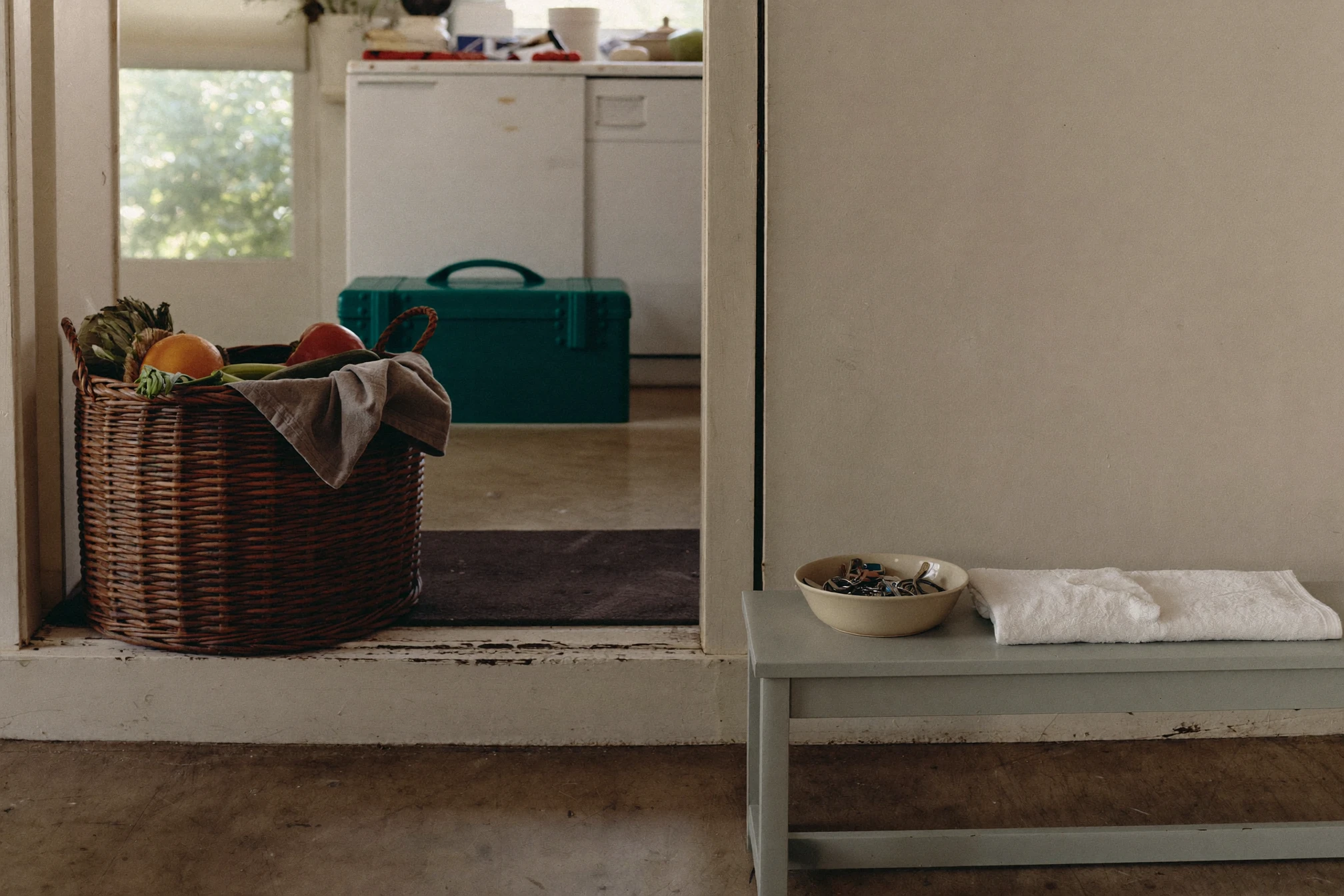 A grocery basket, closed toolbox, folded towels, and keys on a bench between a kitchen and utility room.